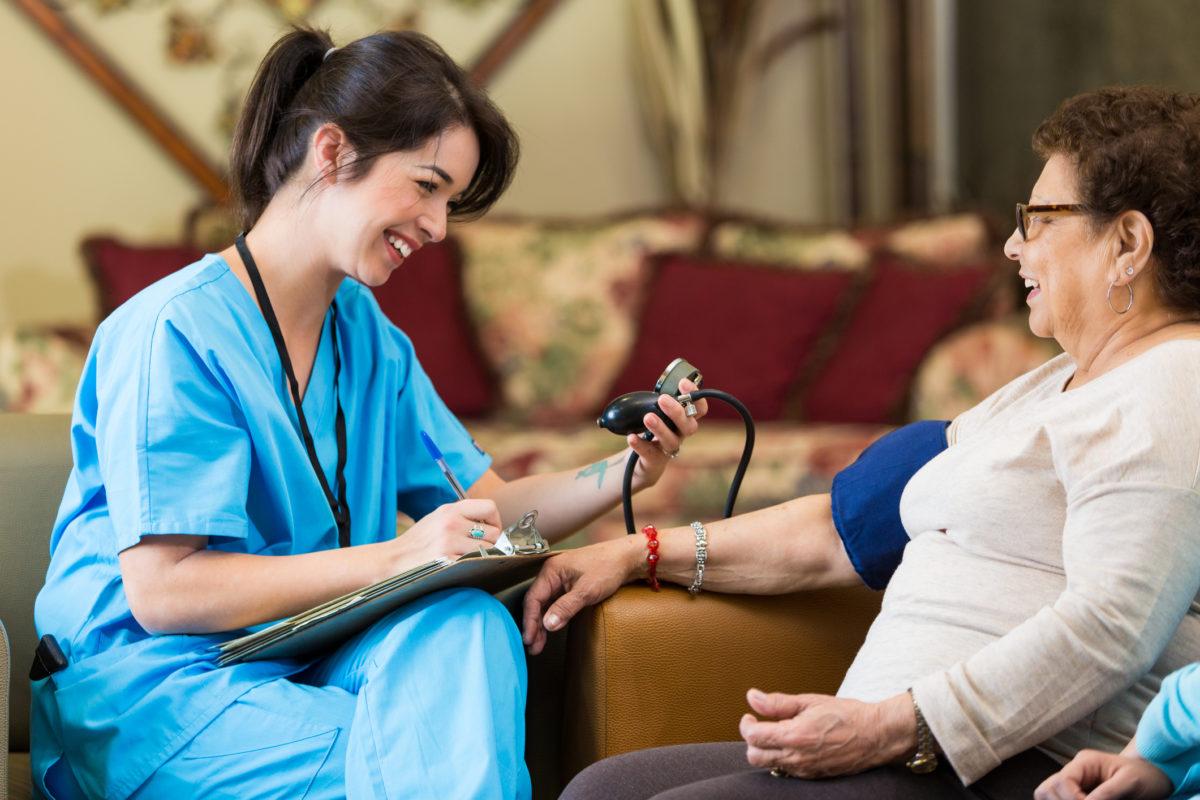 Home healthcare nurse checks senior female patent's blood pressure during home visit. The nurse writes down the patient's blood pressure reading on the patient chart.
